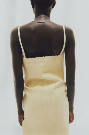 Rear view of a Black woman wearing a yellow slip dress and gold necklace against a seamless grey background.