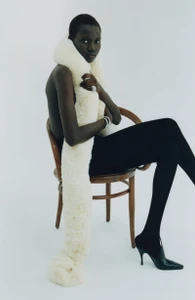 Model seated on a wooden chair wrapped in a long white faux-fur scarf wearing black tights and heels against a white background