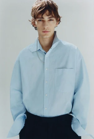 Young man with brown hair wearing a light blue button-down shirt and black trousers against a plain light grey background.