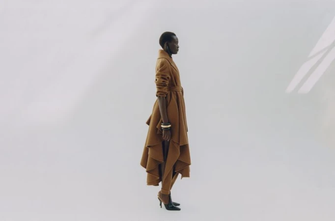 Model standing in profile wearing a structured brown belted coat and high heels against a white backdrop.