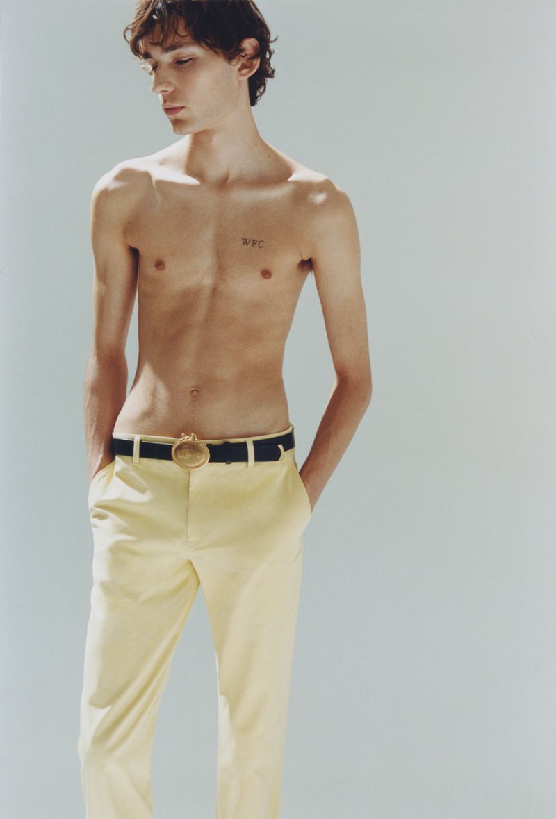 Shirtless young man wearing pale yellow trousers and a dark belt against a plain light grey studio background.