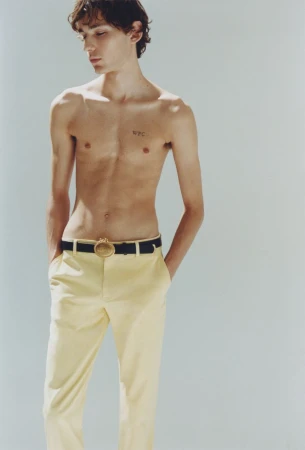 Shirtless young man wearing pale yellow trousers and a dark belt against a plain light grey studio background.
