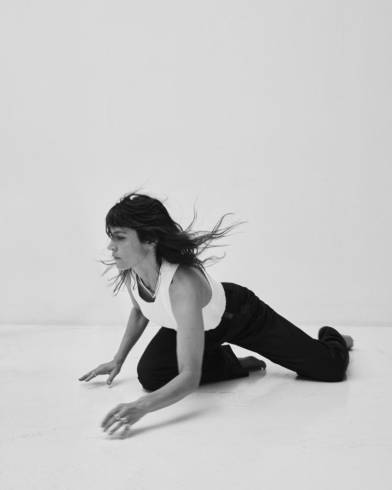 Woman with dark hair blowing in motion wearing a white tank top and black trousers on a white floor