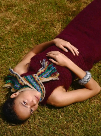 Model lying on grass wearing a burgundy dress and patterned scarf with a grey bracelet.
