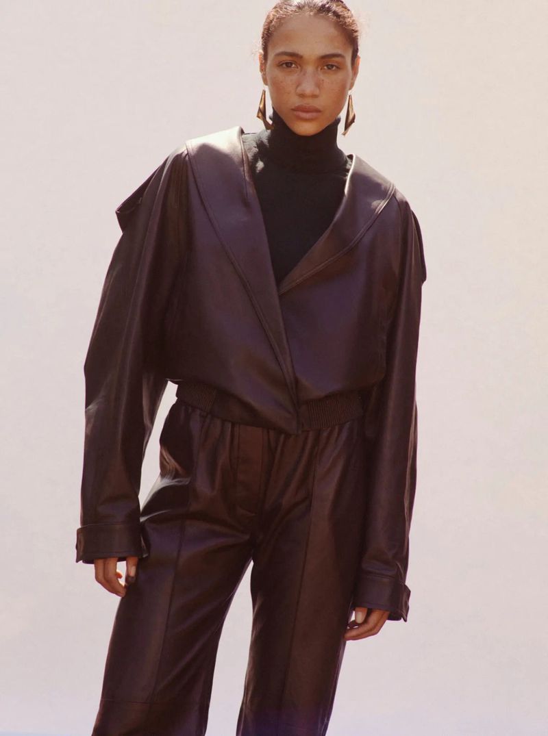 Model wearing a dark brown leather jacket and matching pants with a black turtleneck against a light neutral background.