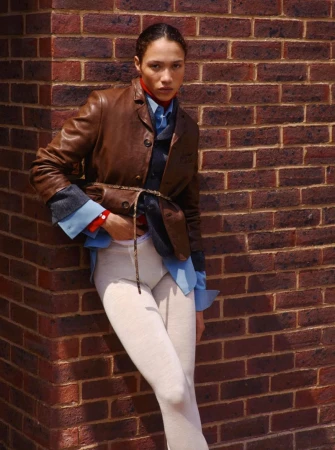 Model leaning against a red brick wall wearing a brown leather jacket, blue shirt, and white leggings.