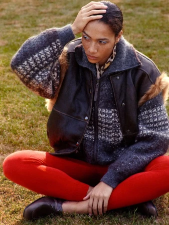Model sitting on grass wearing a textured knit zip-up sweater, dark leather jacket, and red trousers