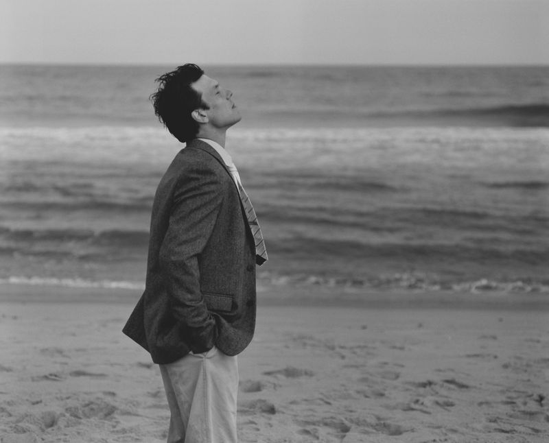 Monochrome portrait of an adult male in a suit looking upwards on a sandy beach with the ocean in the background under daylight.