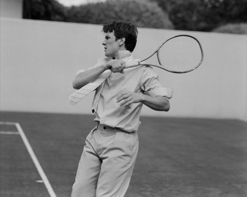 Adult male in collared shirt and tie swings tennis racket on outdoor court, creating a vintage-style image in black and white.