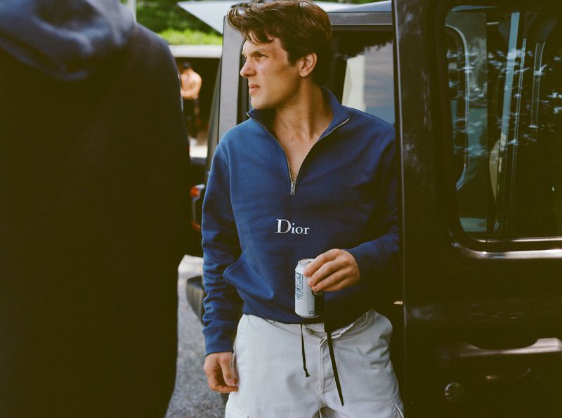 A young man wearing a blue 'Dior' sweatshirt and white shorts is standing near a vehicle and holding a can on a sunny day outdoors.