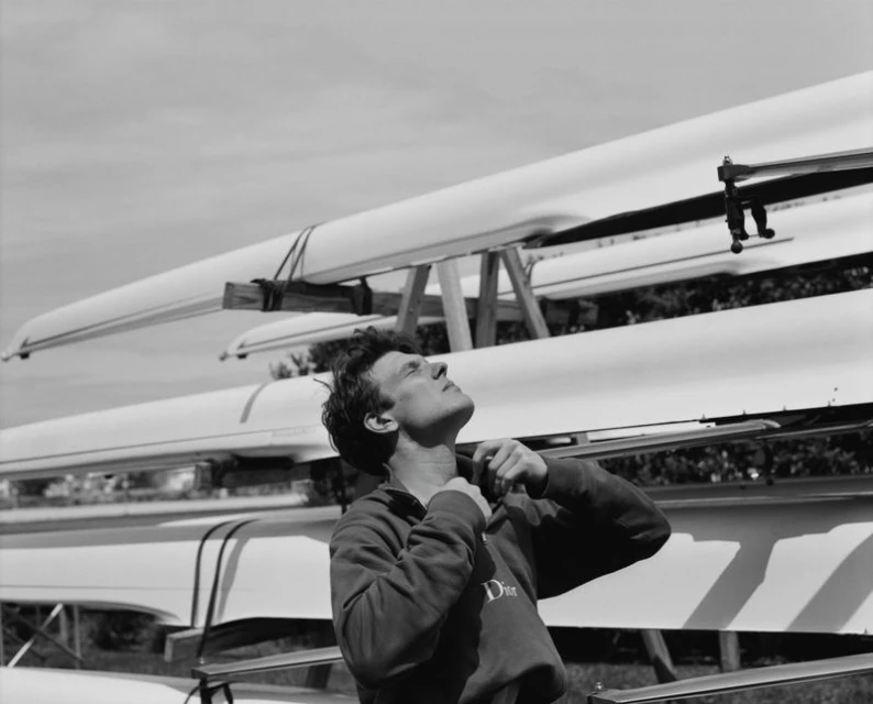 Black and white shot of a fair-skinned adult male standing in front of stacked rowing boats on a sunny day, looking upward.