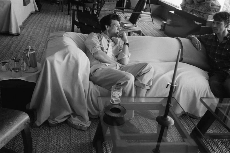 Black and white portrait of two men sitting on a sofa in a sunlit room, one wearing a plaid shirt and the other a suit.