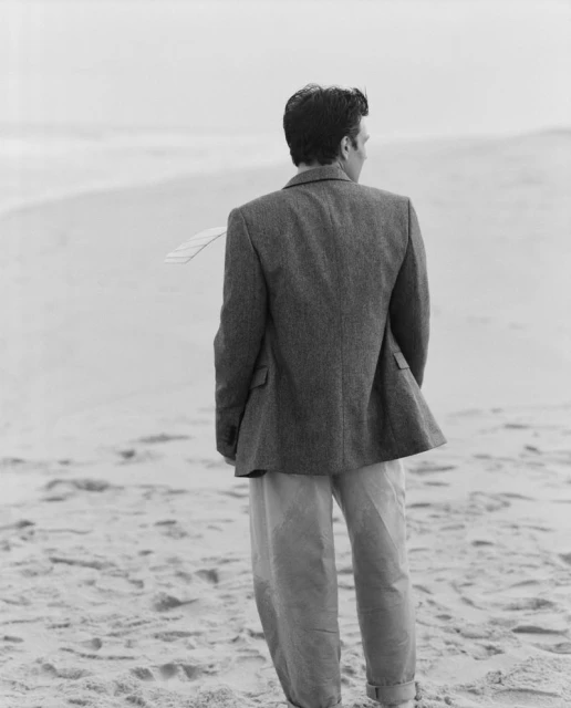 Monochrome portrait of a man facing away, wearing a blazer and trousers, standing on a sandy beach with the ocean in the background.