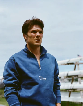 Portrait of a man wearing a blue Dior pullover with a quarter zip standing outdoors on a sunny day with a rack of boats nearby.
