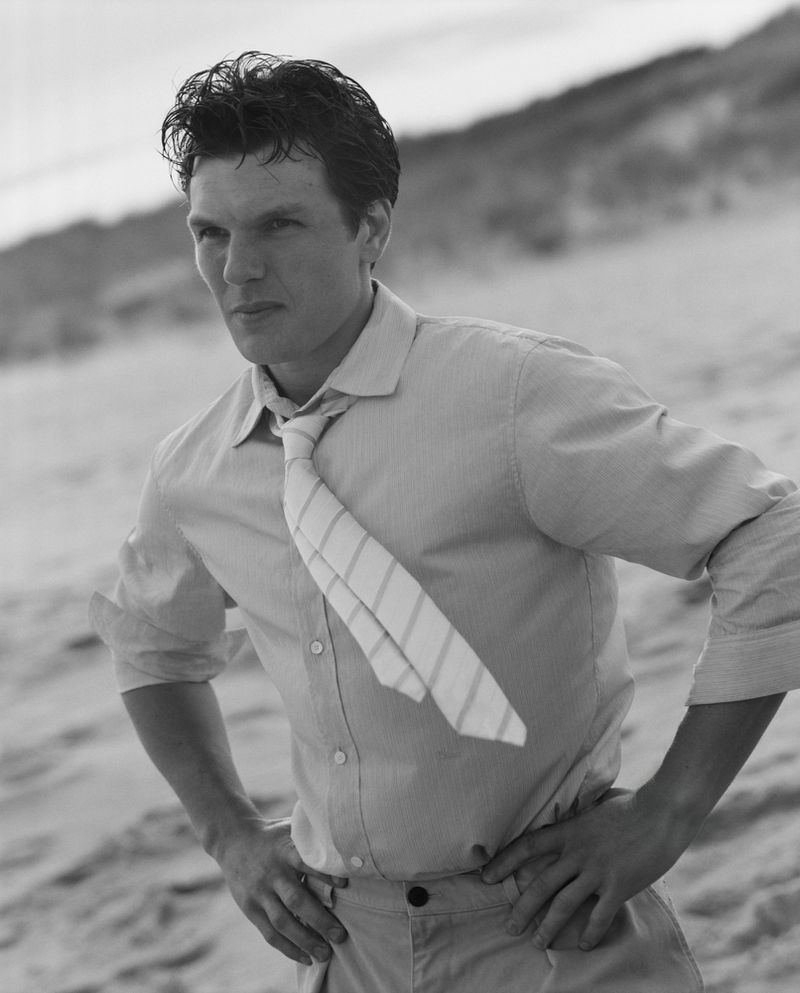 Black and white portrait of a man wearing a shirt and tie, standing on a beach with a sand dune background, medium shot.