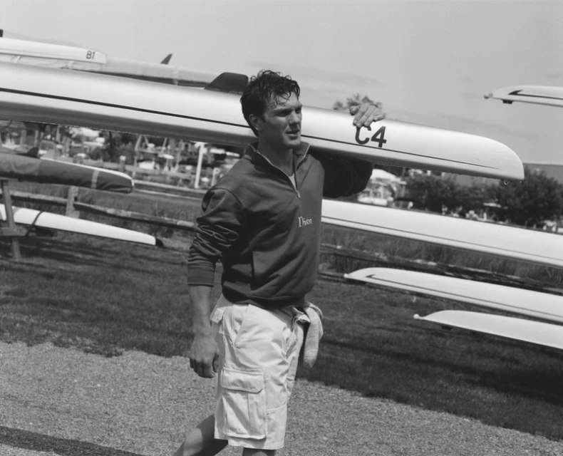 Black and white portrait of adult male carrying a racing scull on his shoulder near a grassy field with multiple sculls.