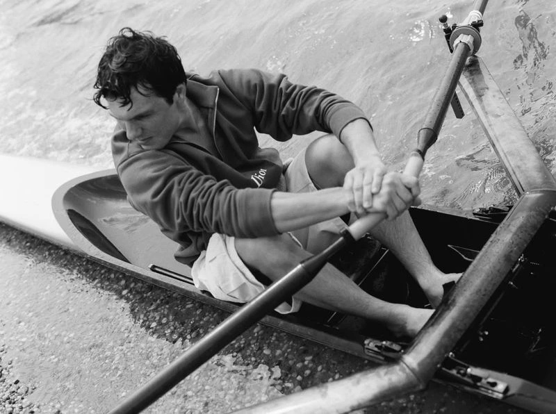 Monochrome image of a male rower in action, seated in a scull with oars in hand, on rippled water, in natural lighting.