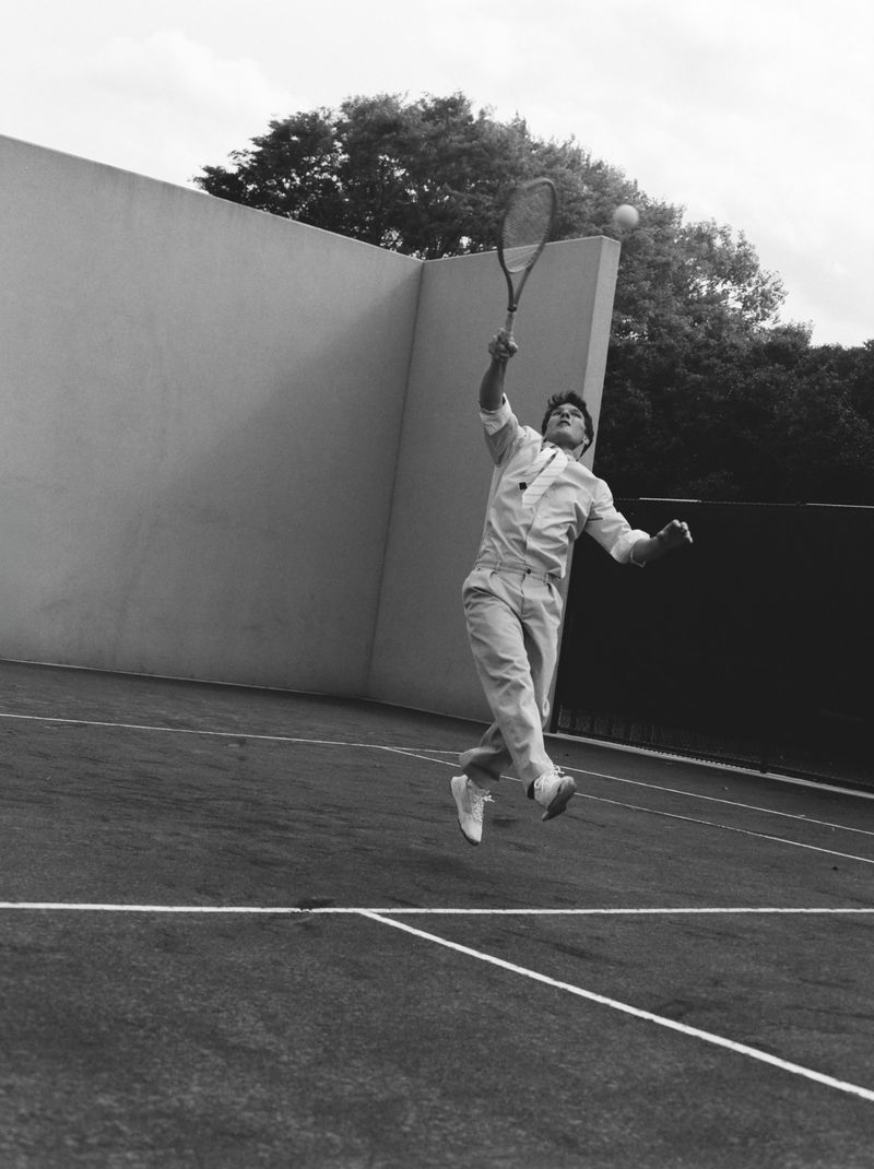 Monochrome image of an adult male tennis player hitting a ball with a racquet on an outdoor tennis court next to a wall and trees.