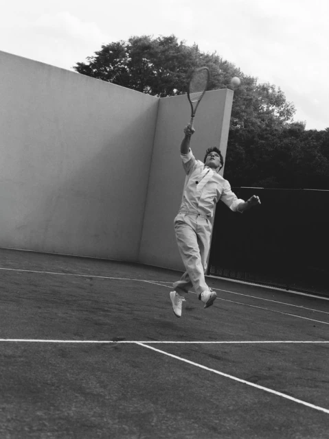 Monochrome image of an adult male tennis player hitting a ball with a racquet on an outdoor tennis court next to a wall and trees.