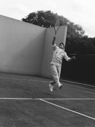 A man in a white collared shirt, tie, and trousers leaps mid-air to hit a tennis ball on an outdoor court in black and white.