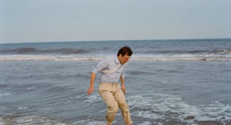 Man wearing a light blue shirt and beige trousers wading in shallow ocean surf on a sandy beach