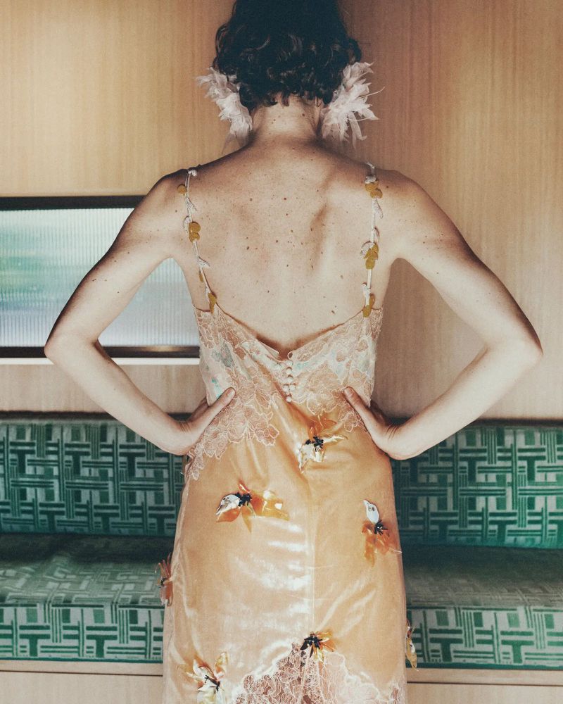 Model seen from behind wearing an embroidered slip dress with feathered hair accessories against a wood-paneled wall