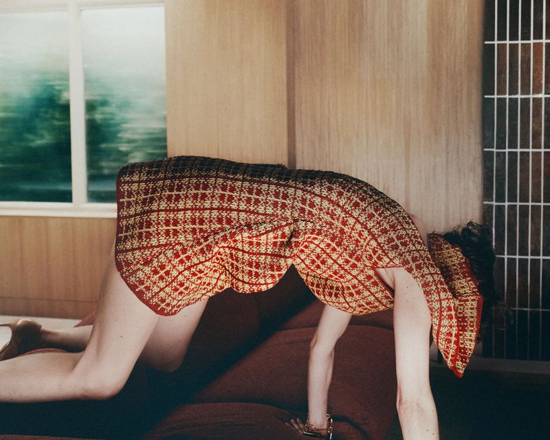 Model wearing a patterned knit dress bending over a sofa in a modern interior setting