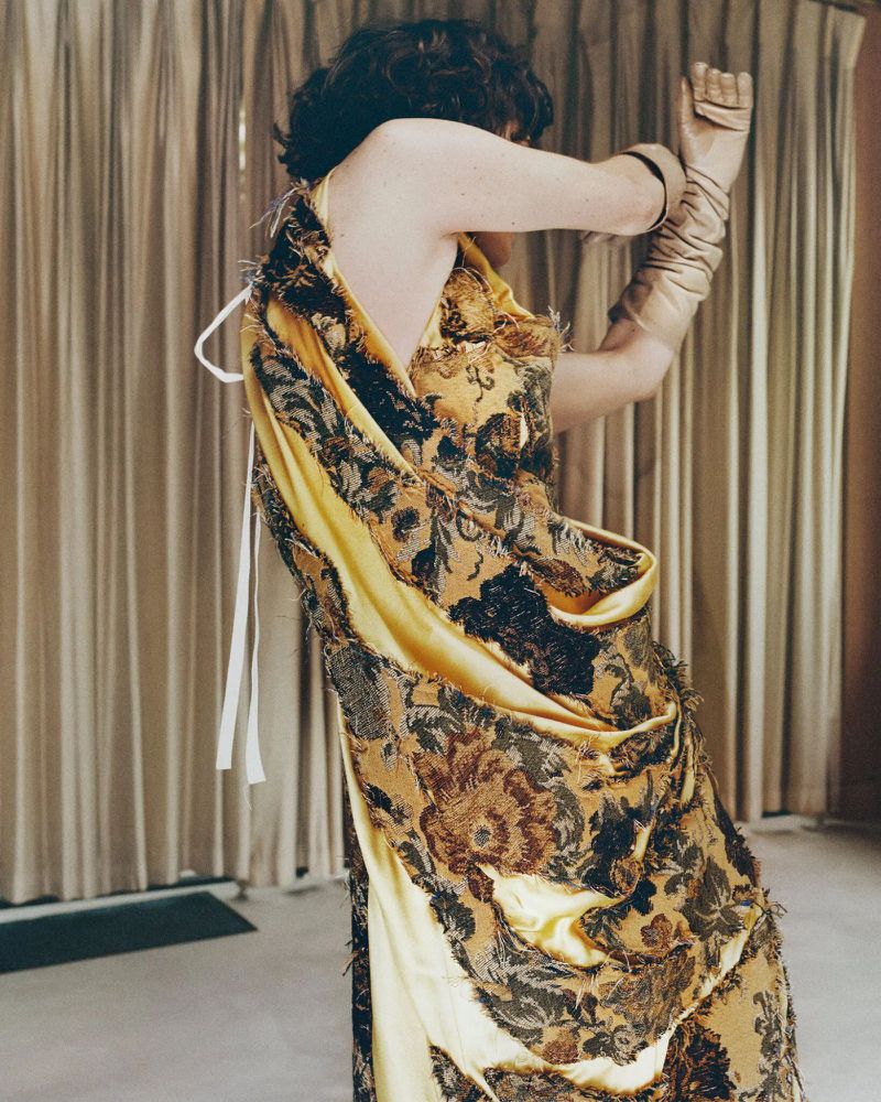 Model in patterned yellow and black dress with matching long gloves posing against pleated curtains