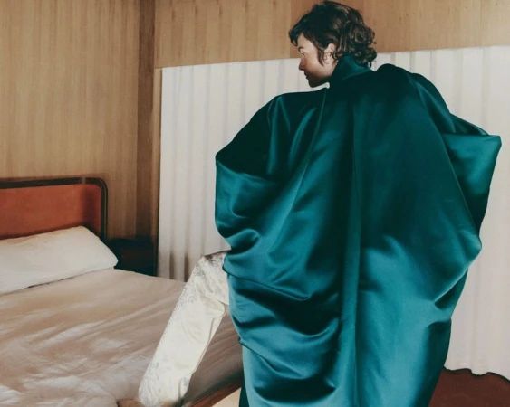 Model in a voluminous dark green silk garment standing beside a bed in a hotel room for T: The New York Times Style Magazine