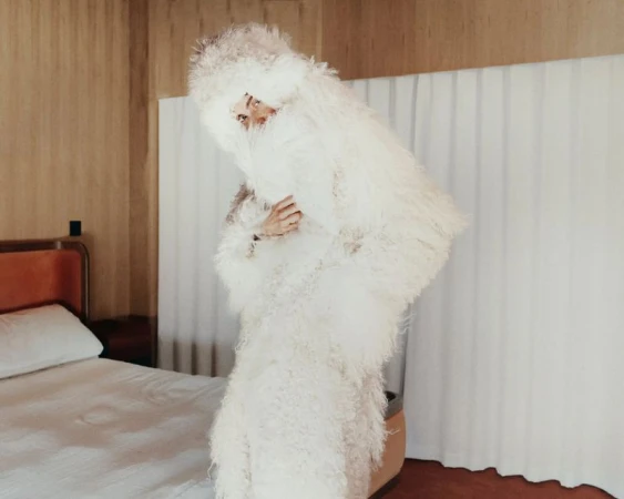 Model wearing a voluminous white shaggy fur coat standing inside a hotel room with a bed