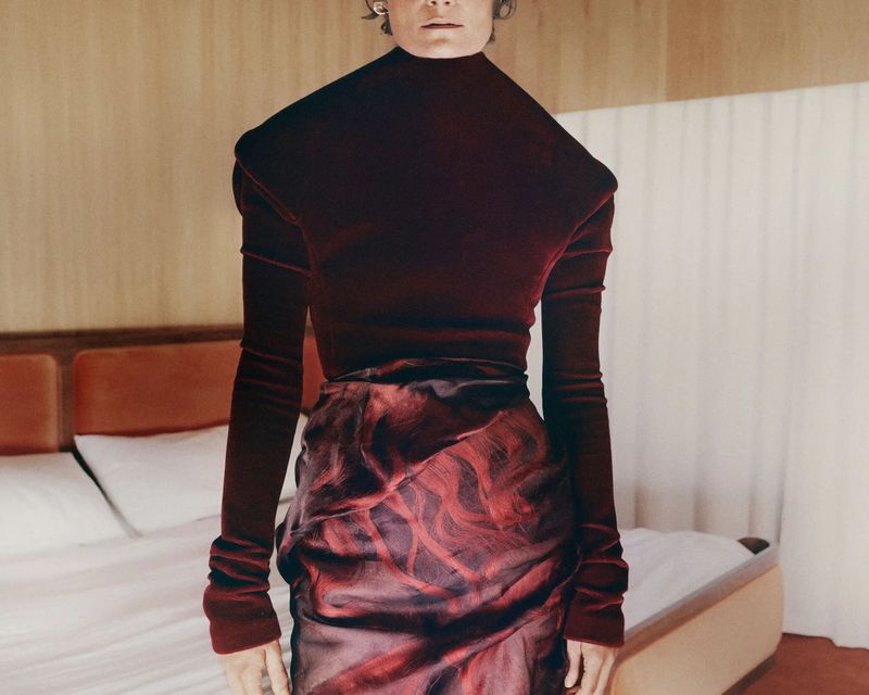 Model wearing a dark burgundy turtleneck top and patterned skirt standing in a bedroom setting