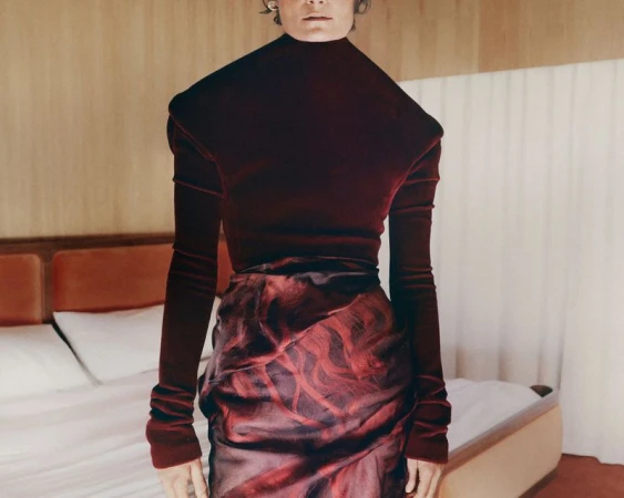 Model wearing a dark burgundy turtleneck top and patterned skirt standing in a bedroom setting