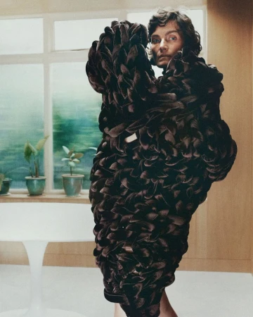 Model wearing a heavily textured dark brown sculptural garment inside a modern interior with potted plants