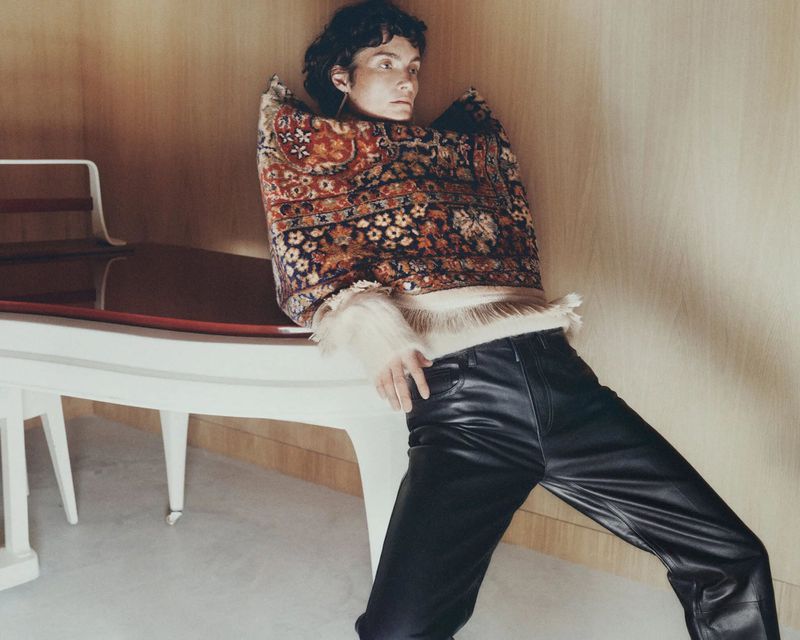 Model wearing a patterned rug as a top and black leather pants leaning against a white piano