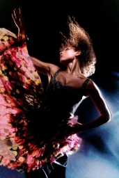 A woman in a dynamic pose wearing a dark patterned dress with her hair swept in motion against a dark background.