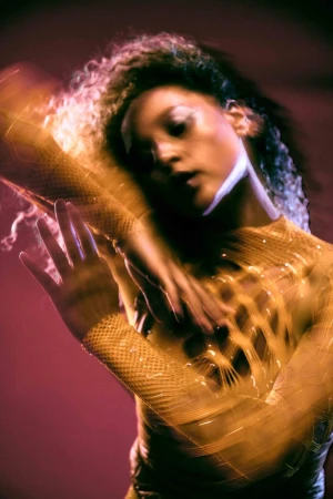Portrait of a woman wearing a textured yellow mesh top with motion blur on her arms against a red and purple background.