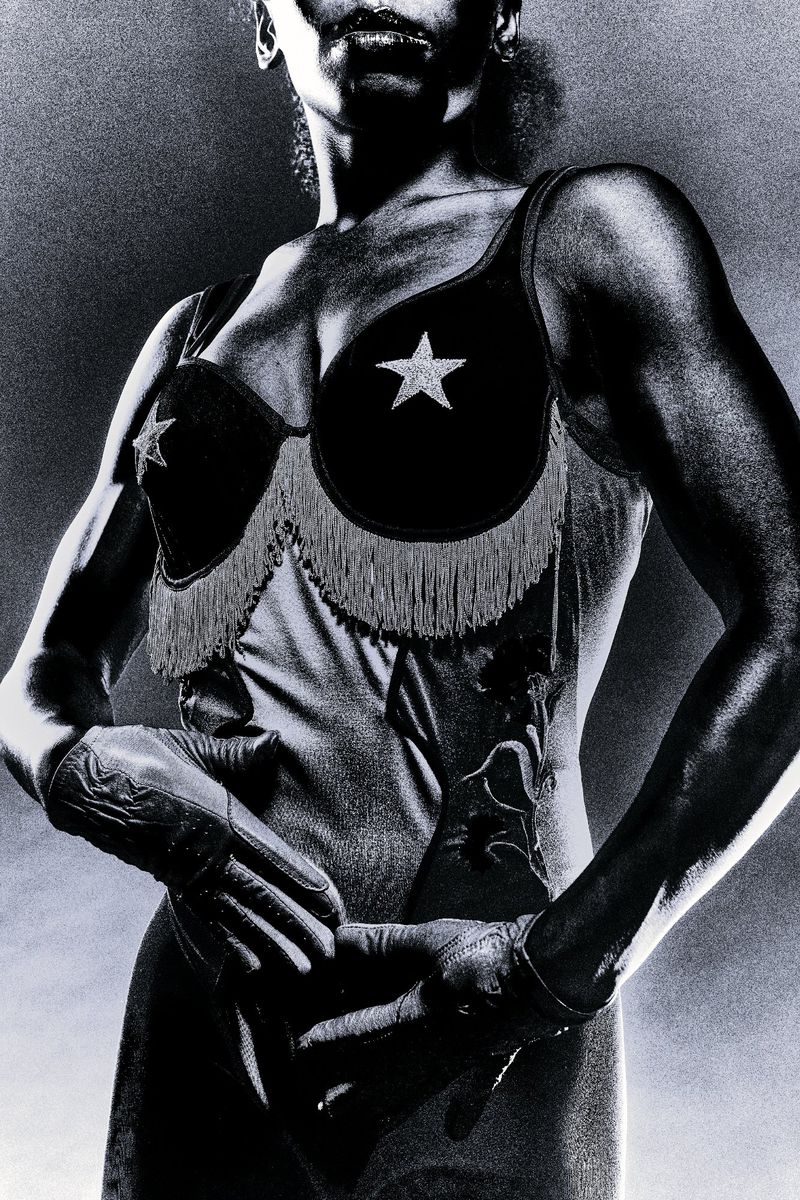Black and white portrait of a muscular woman wearing a star-embellished bustier and gloves against a textured backdrop