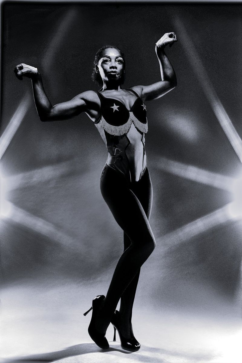 Black and white portrait of a woman posing in a star-patterned bra top and dark high heels against a dramatic lit backdrop.