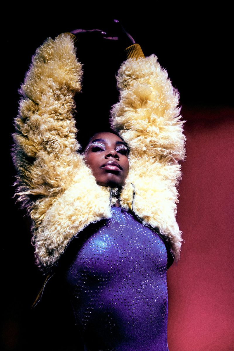 Model with arms raised wearing a textured cream shearling jacket and sparkly purple top for Vingt Sept Magazine by photographer Jacquetta Clark.