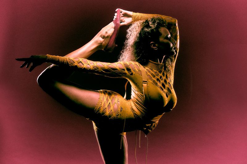 Model in yellow cutout bodysuit performing a dramatic backward flexibility stretch against a dark red background.
