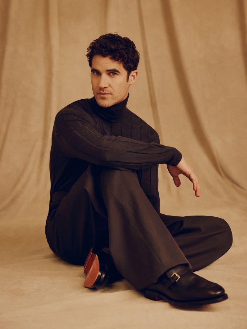 Darren Criss seated on a textured beige backdrop wearing a dark turtleneck and trousers.