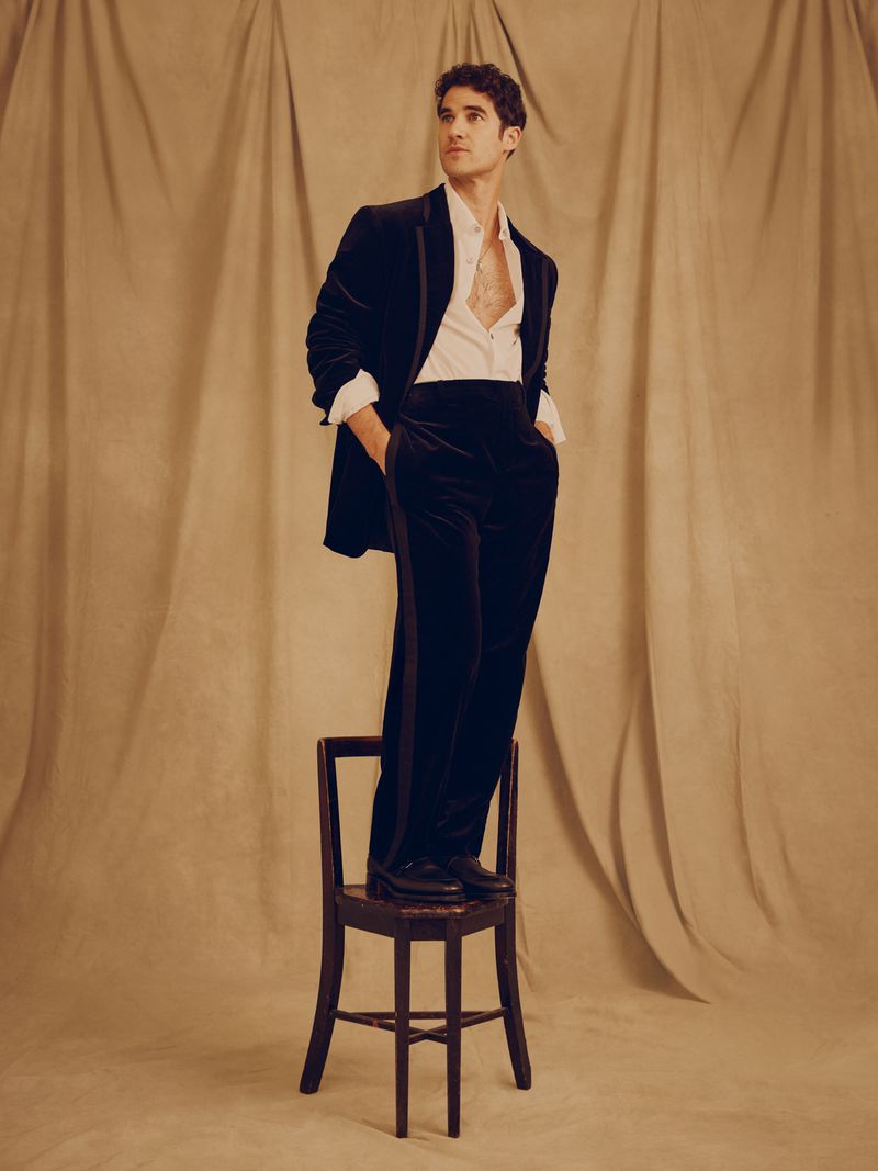 Darren Criss wearing a black velvet suit standing on a wooden chair against a draped tan fabric backdrop