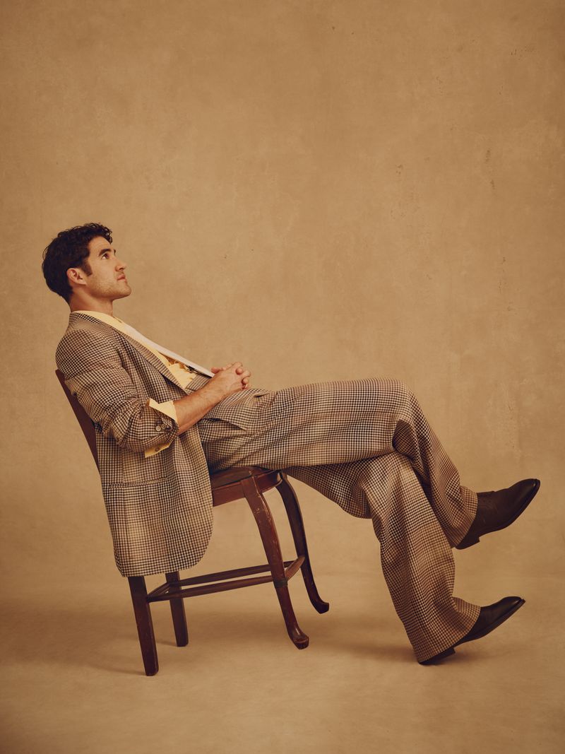Darren Criss seated on a wooden chair wearing a plaid suit jacket and trousers against a neutral textured studio background.