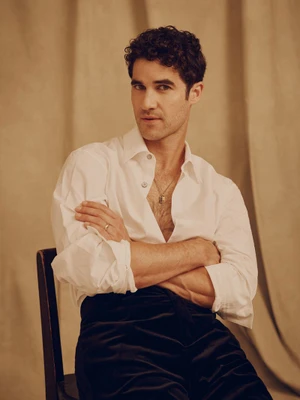 Darren Criss seated with arms crossed wearing a white button-up shirt and dark trousers against a neutral draped backdrop.