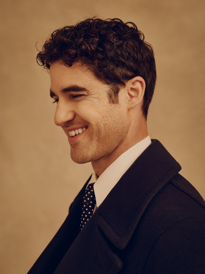 Darren Criss smiling in profile wearing a dark coat and dotted tie against a textured neutral background