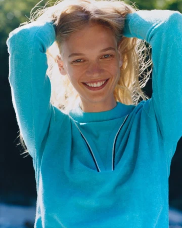 Smiling female model with blonde hair wearing a bright blue sweater holding her hands up behind her head outdoors.
