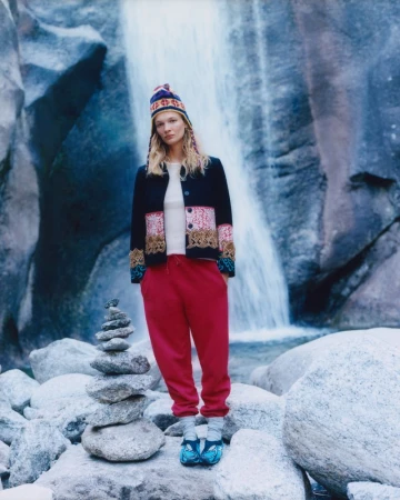 Model Caitlin Soetendal wearing a patterned jacket, red trousers and a beanie hat standing in front of a waterfall