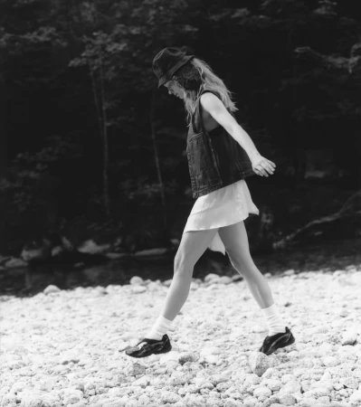 Model Caitlin Soetendal walking along a rocky riverbank wearing a bucket hat, vest, skirt, and sneakers for Pop Magazine.