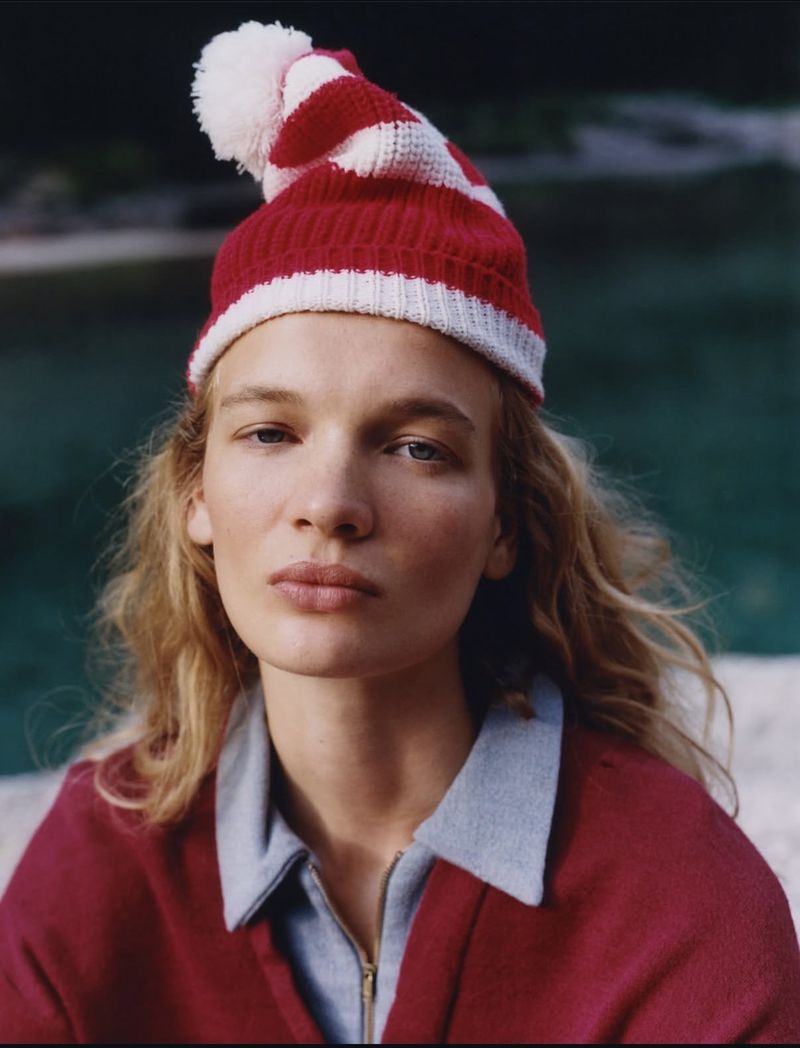 Model Caitlin Soetendal wearing a red and white striped beanie and a maroon zip-up top against a dark water background.