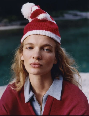 Model Caitlin Soetendal wearing a red and white striped beanie and a maroon zip-up top against a dark water background.
